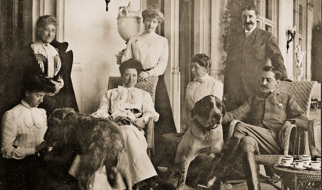 Archival photo of a group of men and women, plus two large dogs, having afternoon tea at Biltmore House