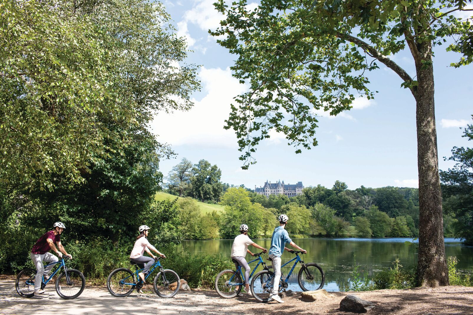 A family enjoys a bike ride during their Biltmore visit. They are paused in front of the Lagoon with a view of Biltmore House in the distance.