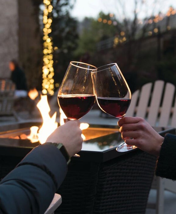 Wine glasses clink in celebration before lit outdoor fire pit