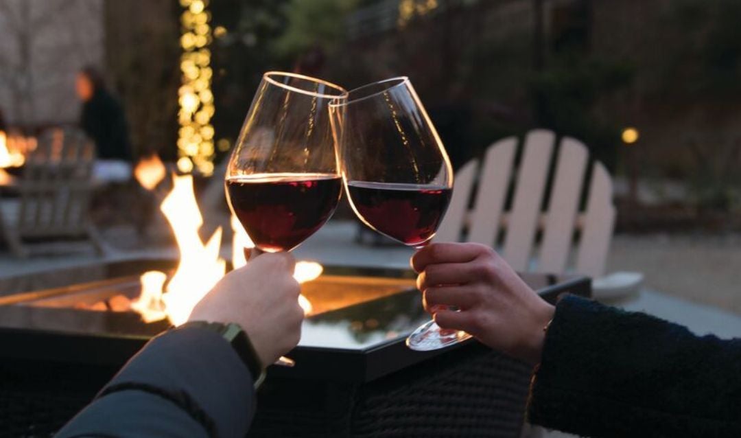 Two people toasting with glasses of red wine in front of a firepit.