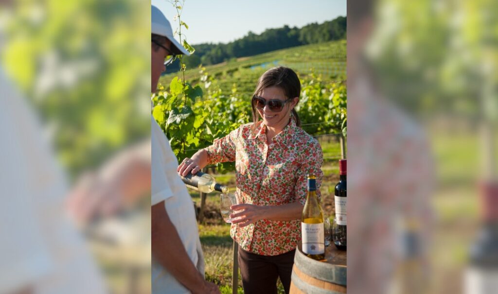 A woman in sunglasses pours wine in a vineyard.