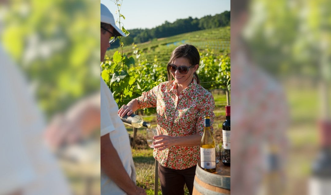 A woman in sunglasses pours wine in a vineyard.
