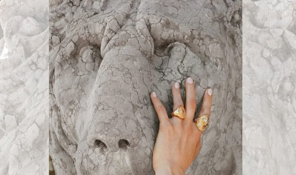 A woman's hand adorned with gold rings touches the face of a marble lion in front of Biltmore House.