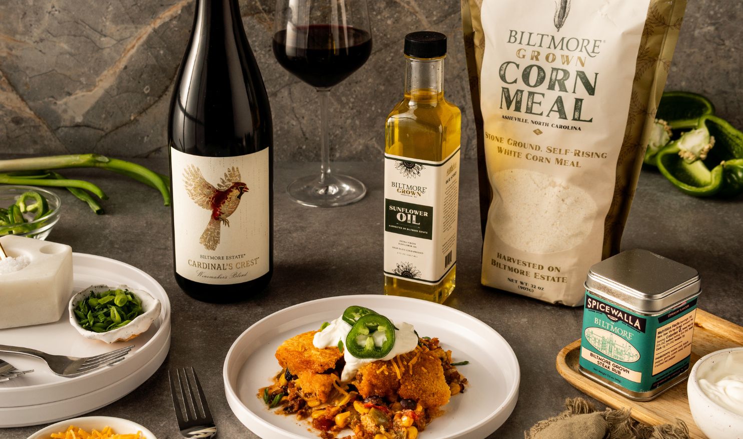 Biltmore Grown Harvest Chili Cornbread on a plate with a glass and bottle of Biltmore Estate Cardinal's Crest red wine, Spicewalla rub, Biltmore Grown Sunflower Oil, Biltmore Grown Cornmeal Mix, and chili toppings.