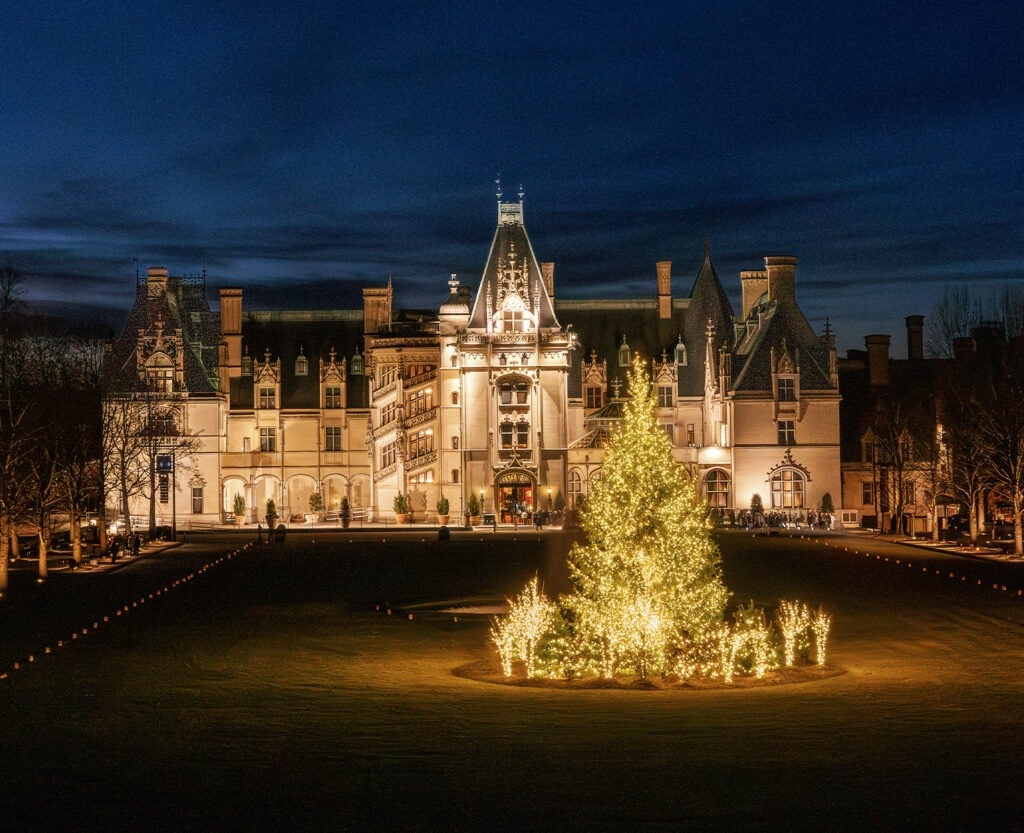 Biltmore House facade and front lawn trees cast in warm evening glow from christmas lights